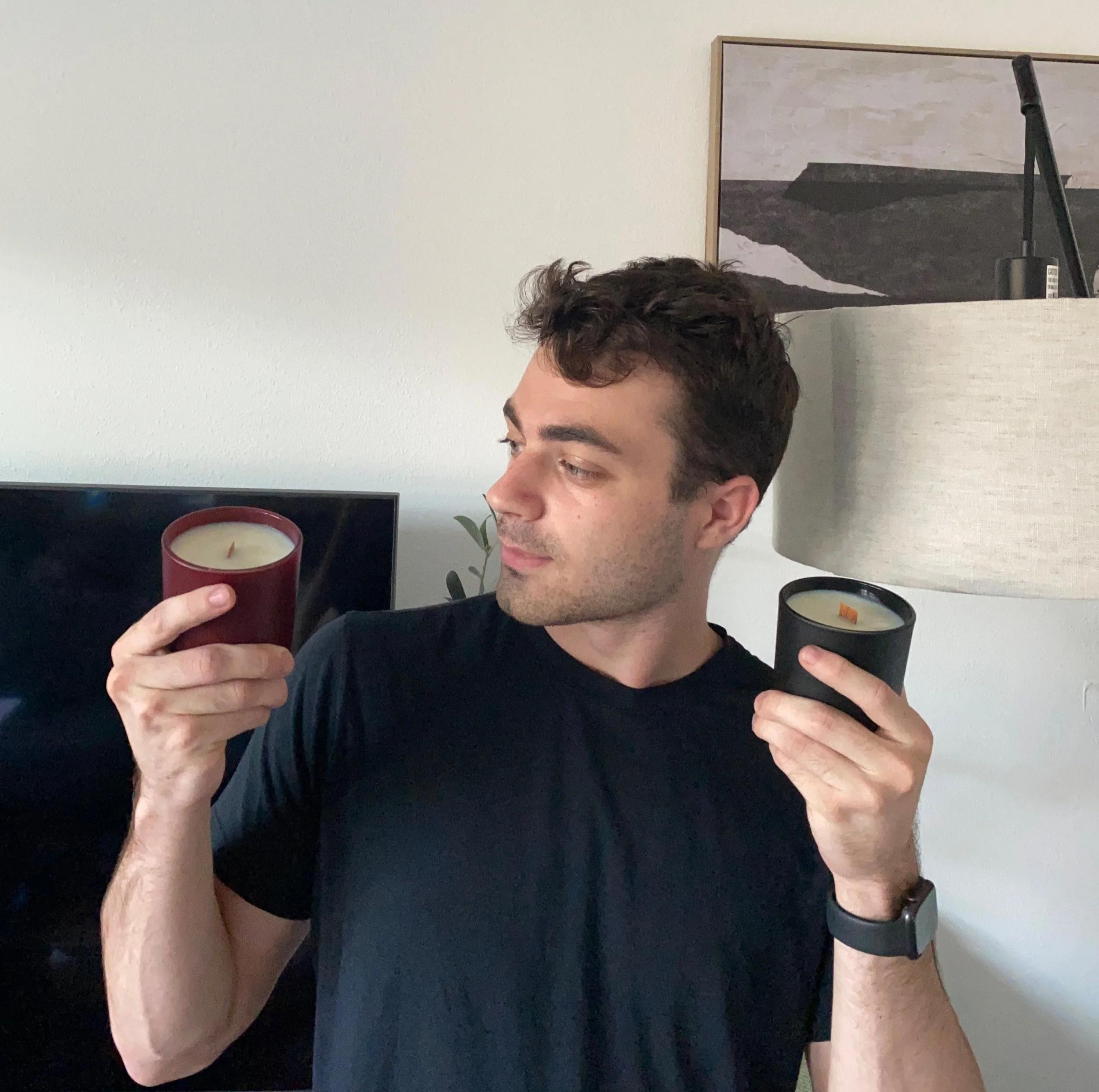 A young man with curly brown hair and light skin holding two candles, one red and one black, while looking at them.