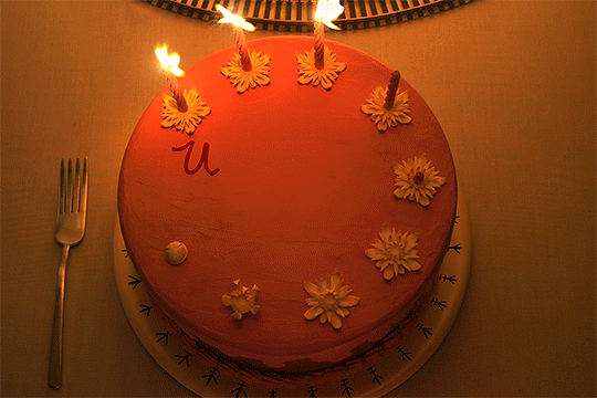 A pink birthday cake decorated with yellow flowers and candles, with a fork on the left side.