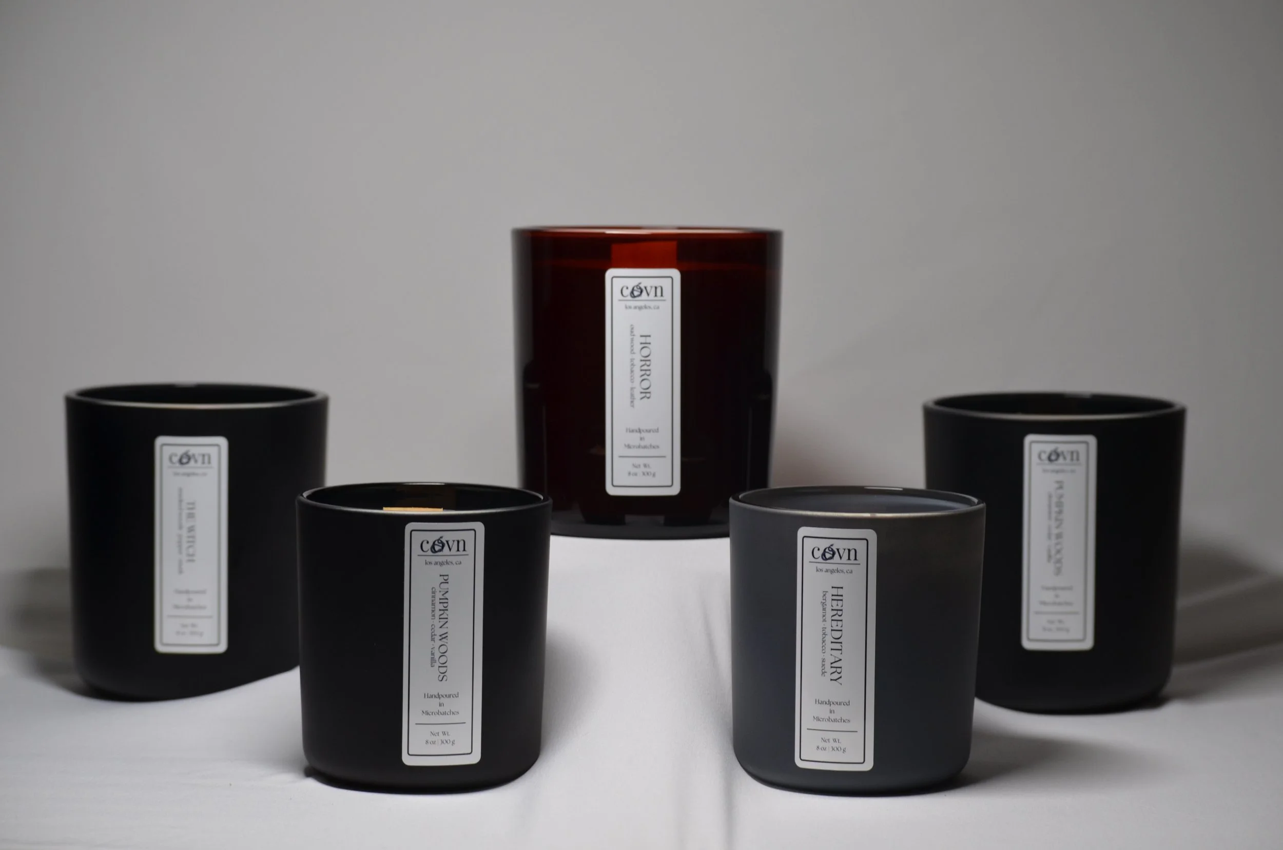Black and dark red candles with labels from the Coven brand arranged on a white surface against a plain background.