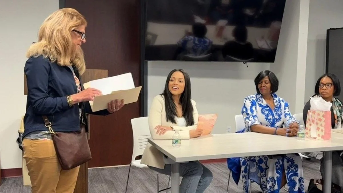 A woman with blonde hair and glasses stands reading from a folder in a conference room. Three women sit at a table listening; one is smiling and holding a folder, the second is in a blue and white dress with folded hands, and the third in glasses and a scarf has a gift bag and water bottles in front of her.