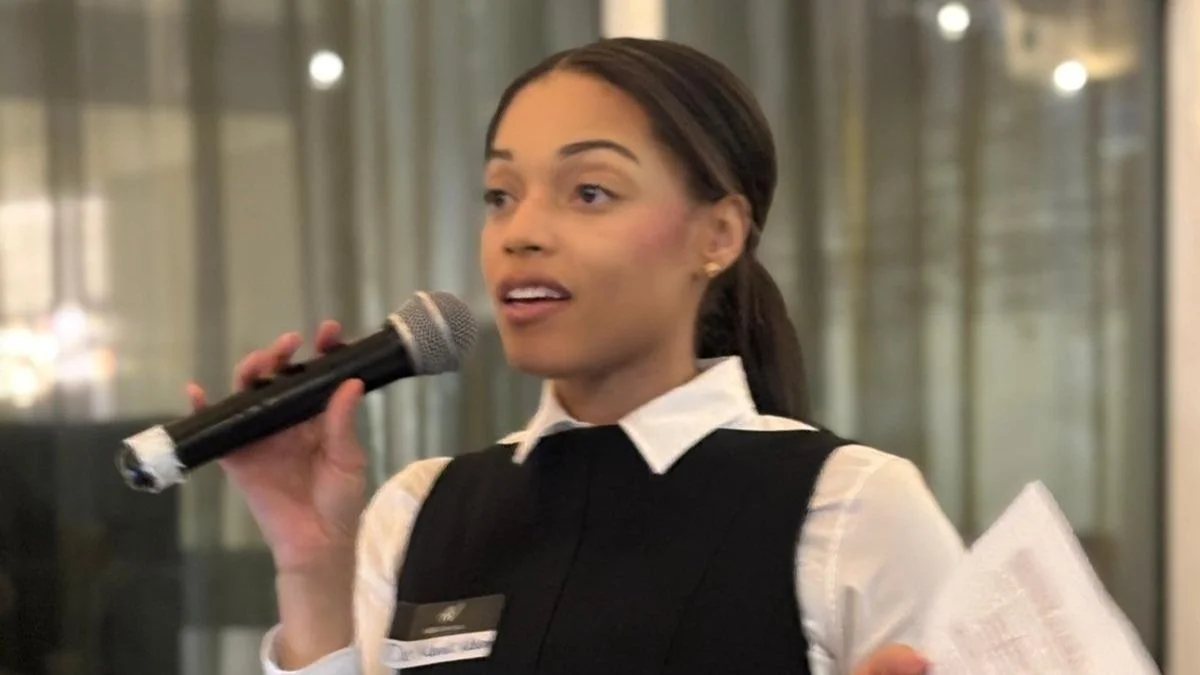 A woman holding a microphone speaking at an indoor event, wearing a formal black vest over a white shirt, with a name tag on her chest, and holding papers in her other hand.