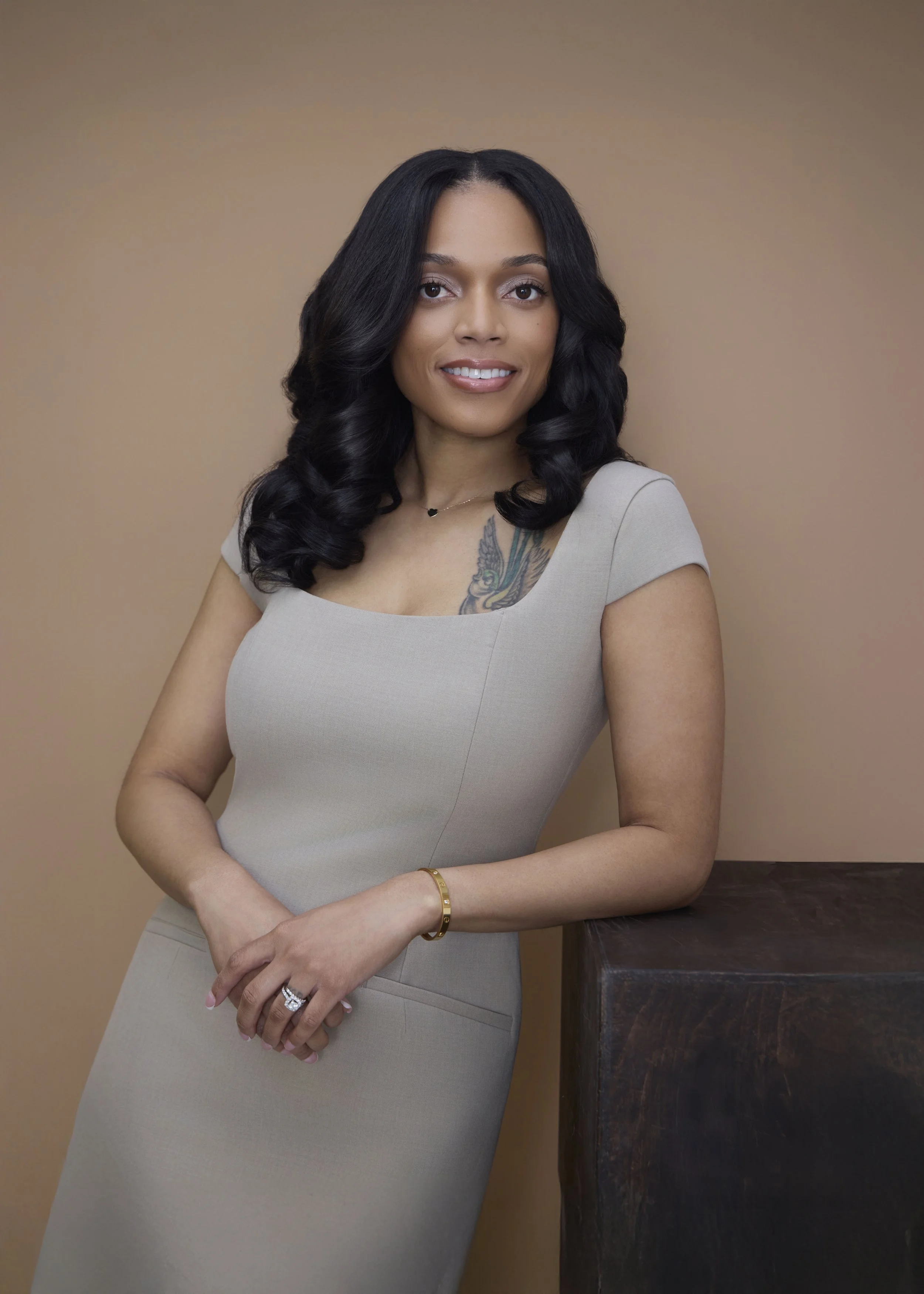 A woman with dark, curly hair, wearing a beige dress, standing next to a wooden table against a neutral background.