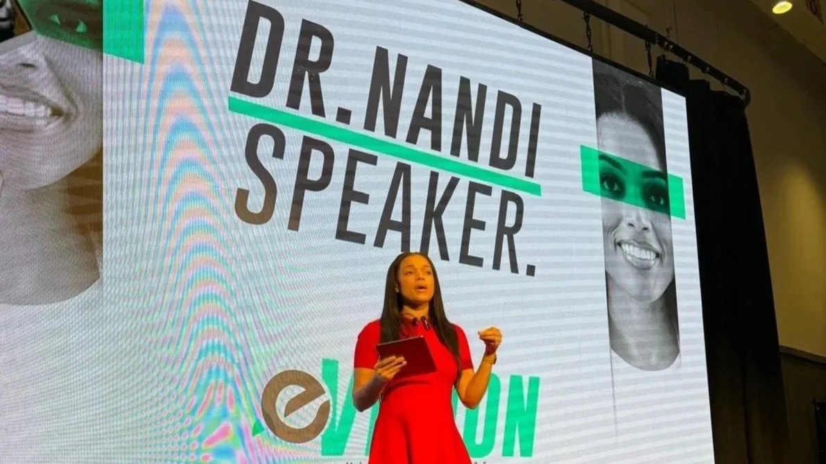 A woman in a red dress speaking on stage with a large screen behind her displaying text that says "Dr. Nandi Speaker" and various images of smiling faces.