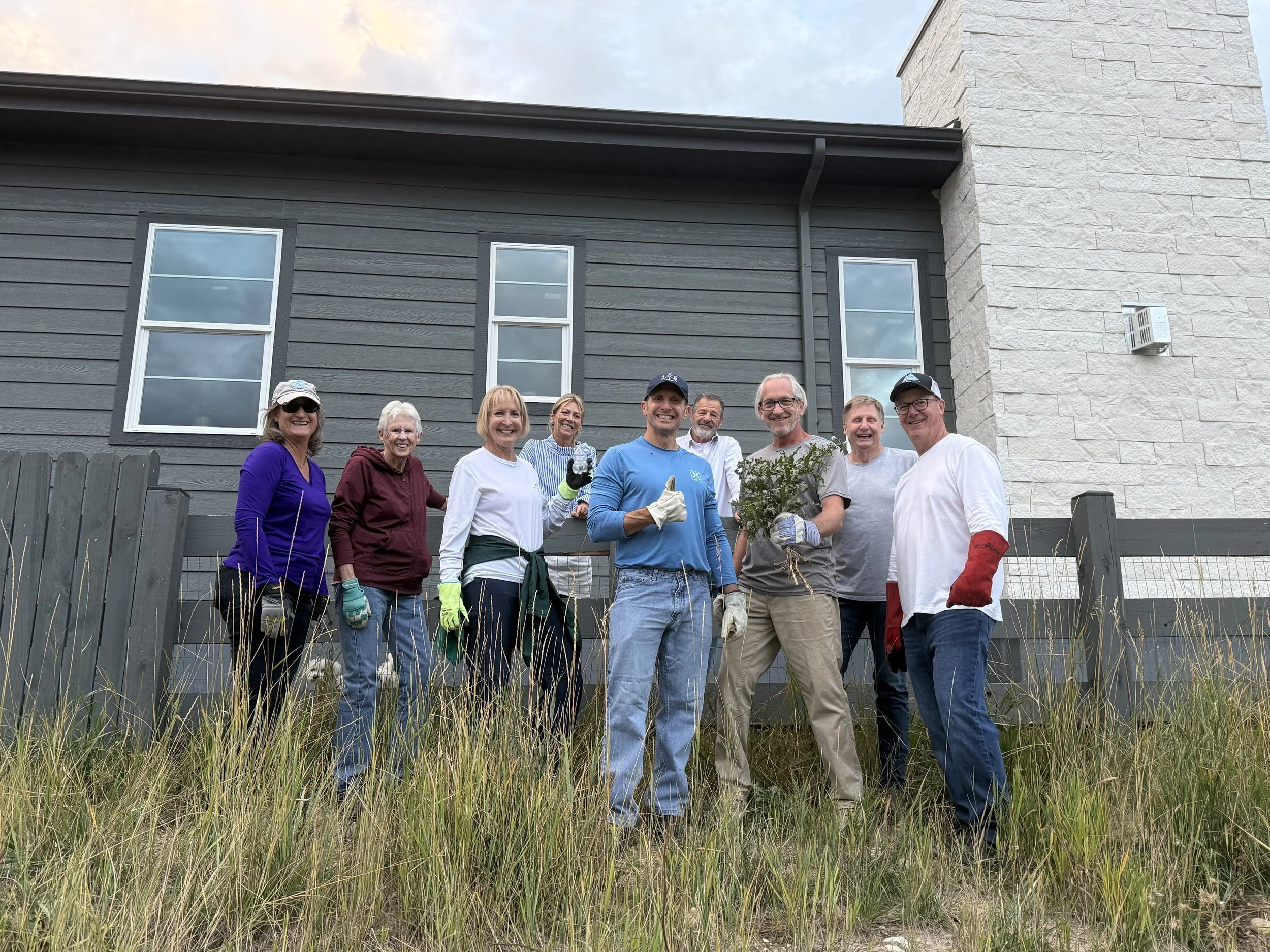 Group of people standing outdoors in front of a house, holding gardening tools and a potted plant, smiling for the photo.