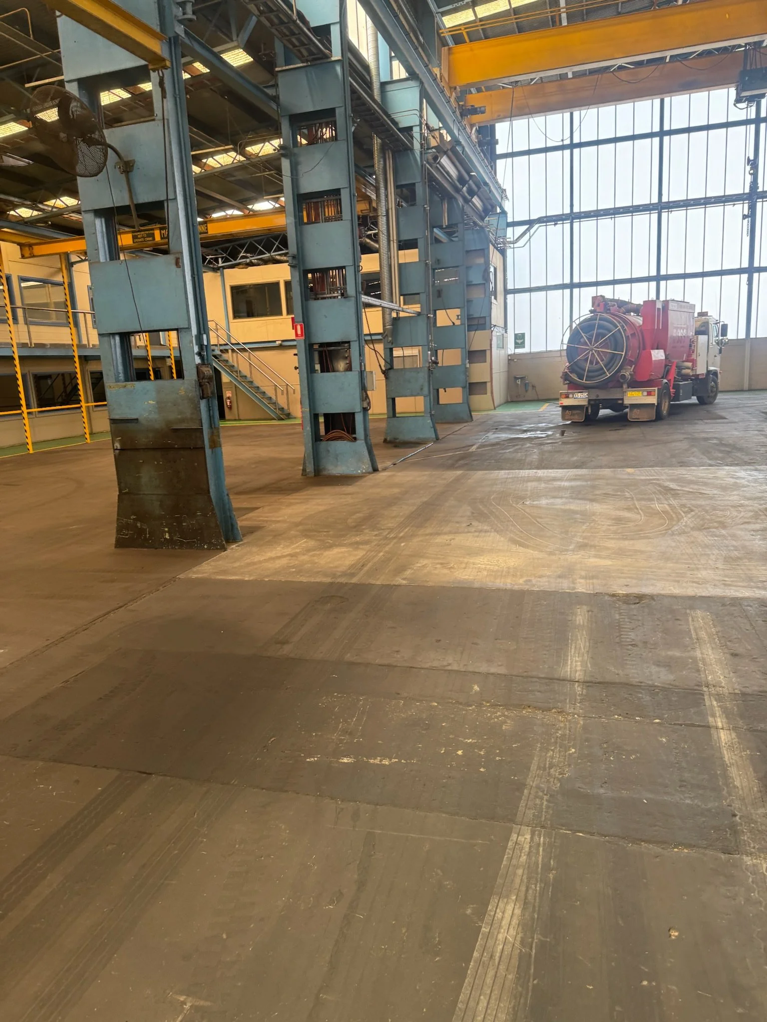 Interior of a large industrial or warehouse building with a high ceiling, large windows, and a concrete floor. There is a red fire truck or equipment vehicle parked inside, with yellow overhead cranes and metal structural elements visible.
