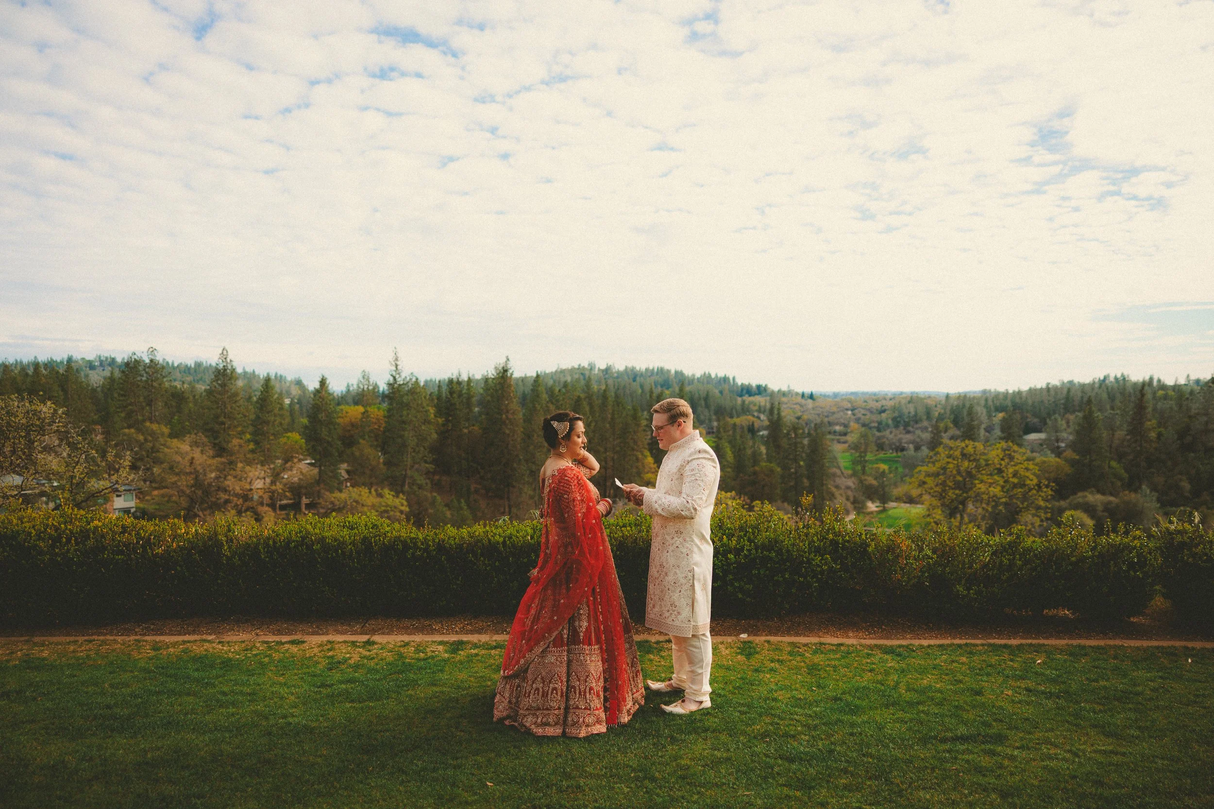 A couple dressed in traditional Indian attire stands outdoors, exchanging vows or rings against a scenic landscape with trees and a cloudy sky.