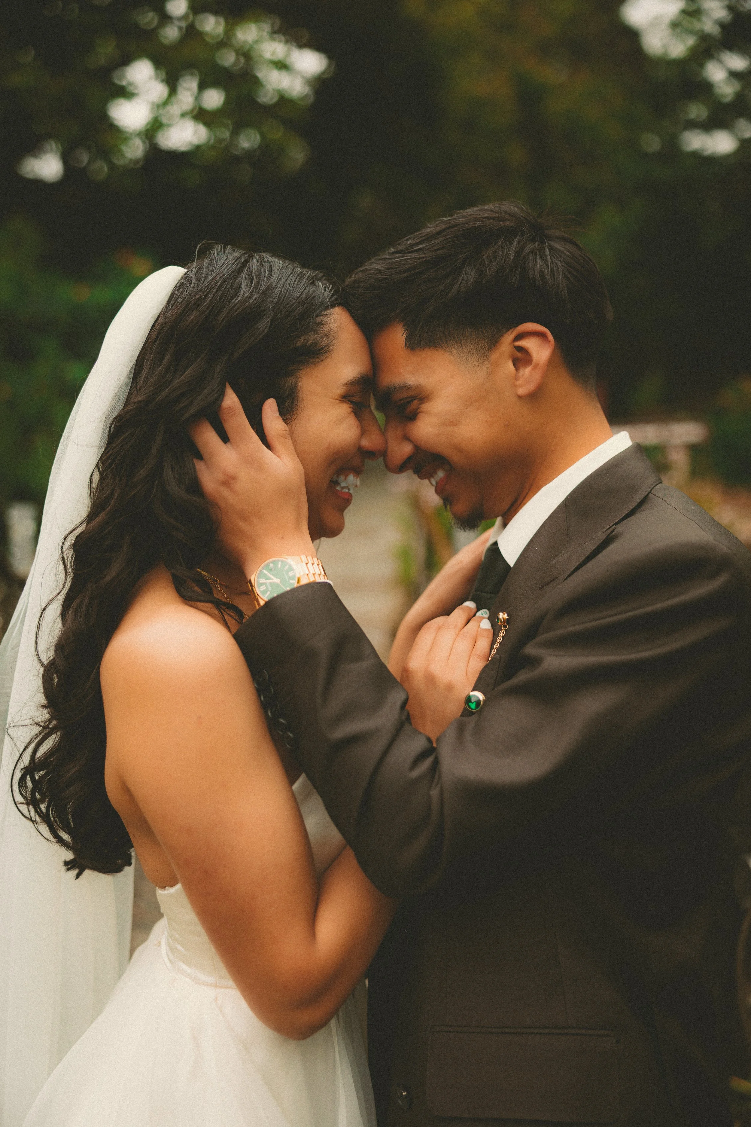 A bride and groom are holding each other with their foreheads touching and eyes closed, sharing a joyful moment outdoors. The bride has long, dark, curly hair, wears a white wedding dress and a veil. The groom is dressed in a dark suit with a white shirt and tie, and has short dark hair.