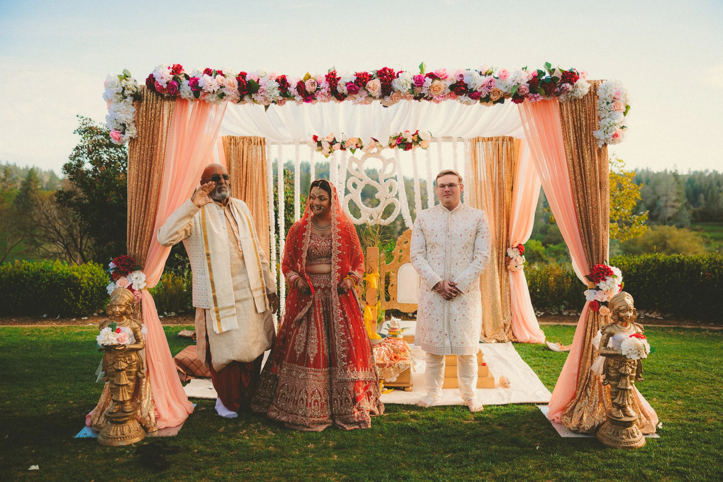 A wedding ceremony with a bride and groom standing under a decorated arch with floral arrangements, and an officiant saluting on a grassy outdoor setting.