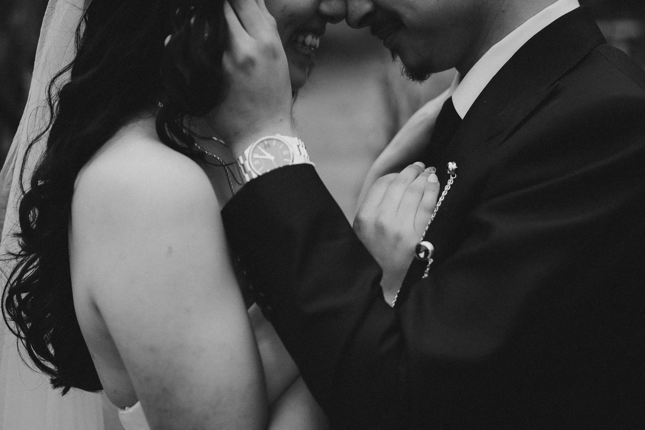 Close-up of a couple touching foreheads, smiling and sharing an intimate moment. The woman has long hair and is wearing a watch, and the man is dressed in a suit.