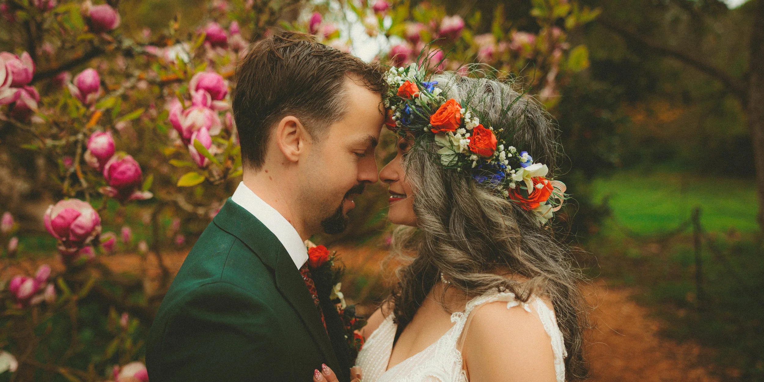 A man and woman with closed eyes touching foreheads, with the woman wearing a floral crown, in an outdoor garden with blooming pink flowers.