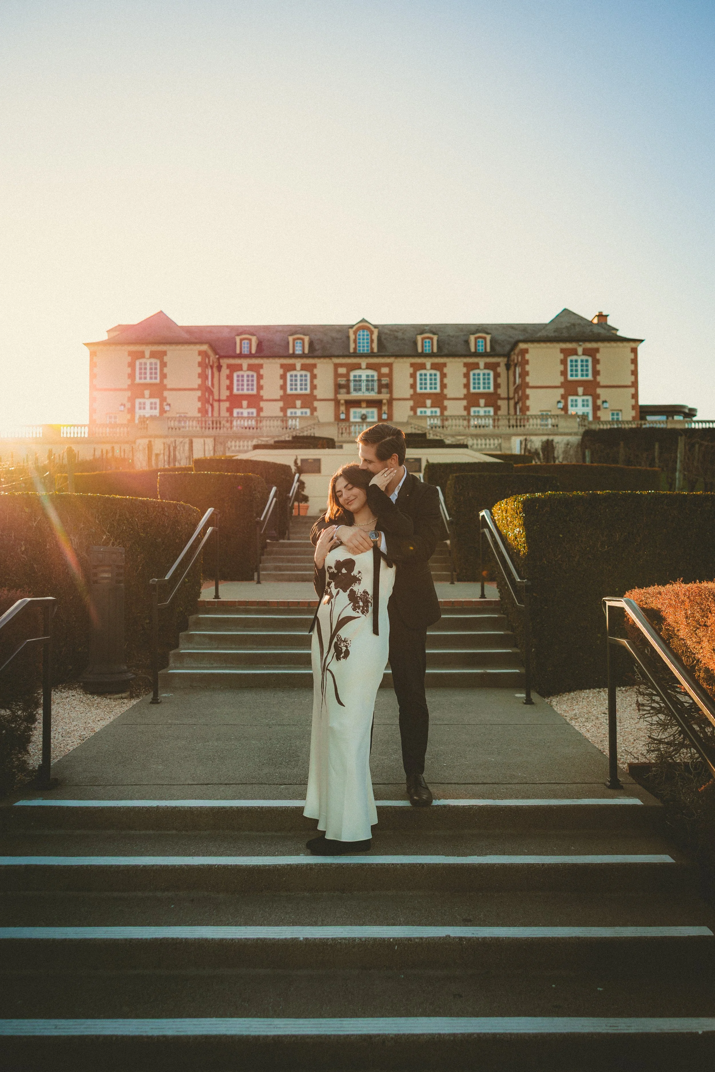 A romantic couple hugging on steps outside a large building during sunset, with a clear sky in the background.