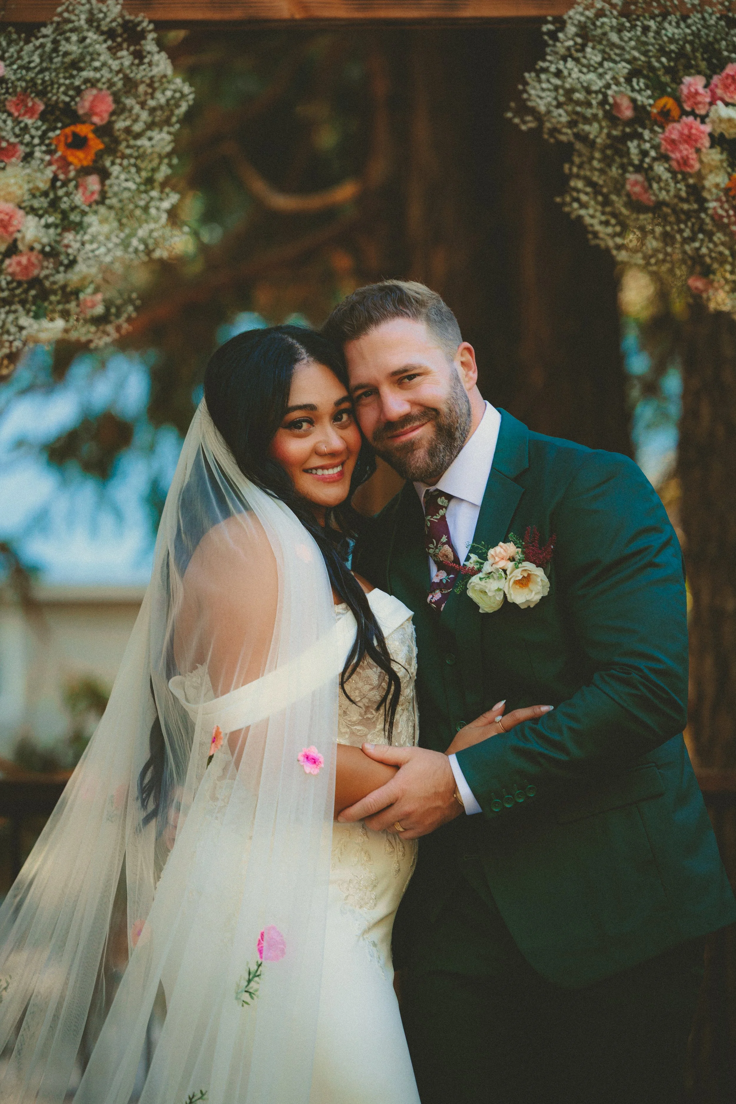 A newlywed couple smiling, embracing, in wedding attire, standing outdoors with flower arrangements and a wooden background.