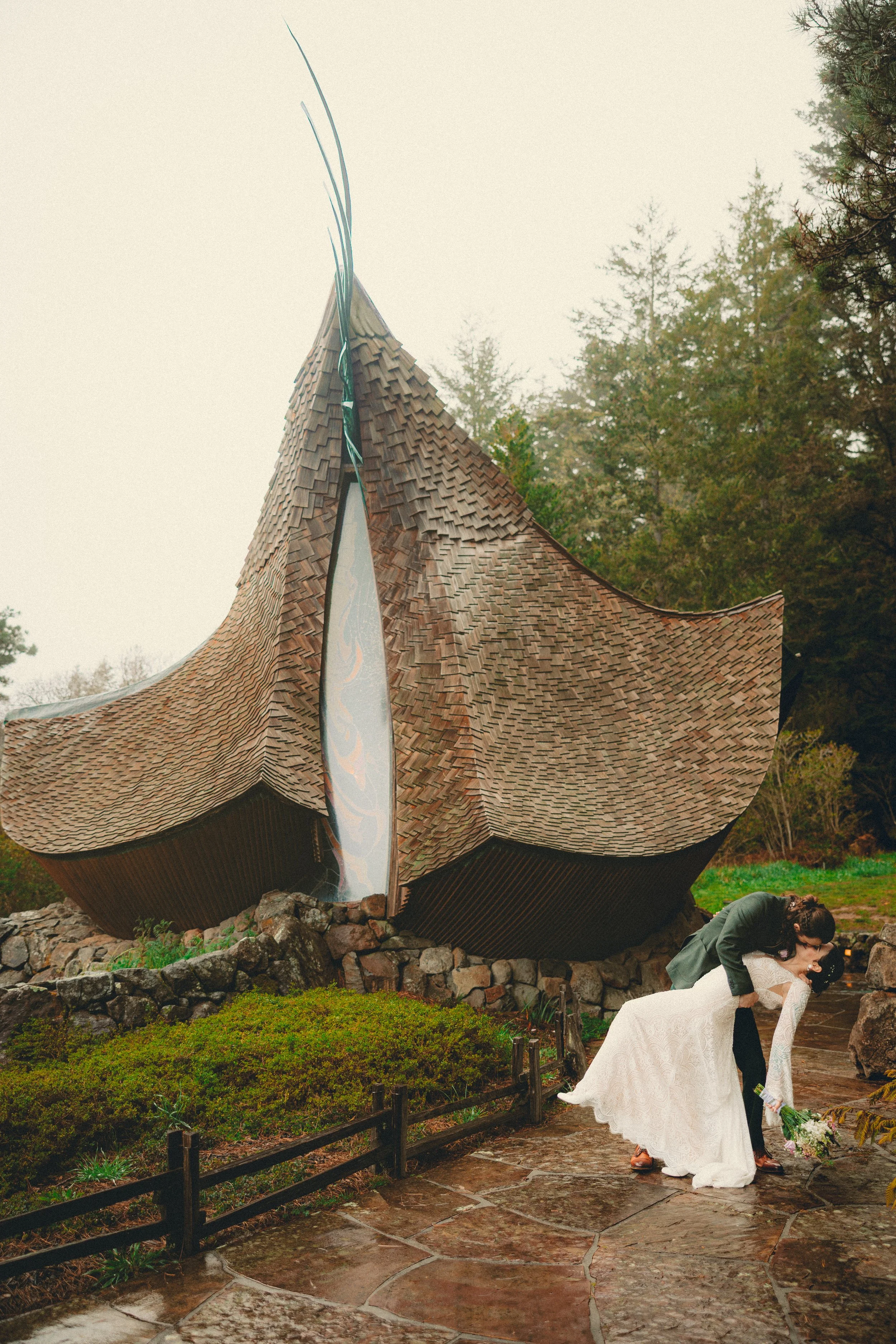 A woman in a wedding dress receiving a kiss from a man in a suit during a wedding ceremony outdoors in front of a unique, architecturally distinctive wooden building with a curved roof and wooden shingles. The scene is on a stone pathway with greenery and trees in the background.