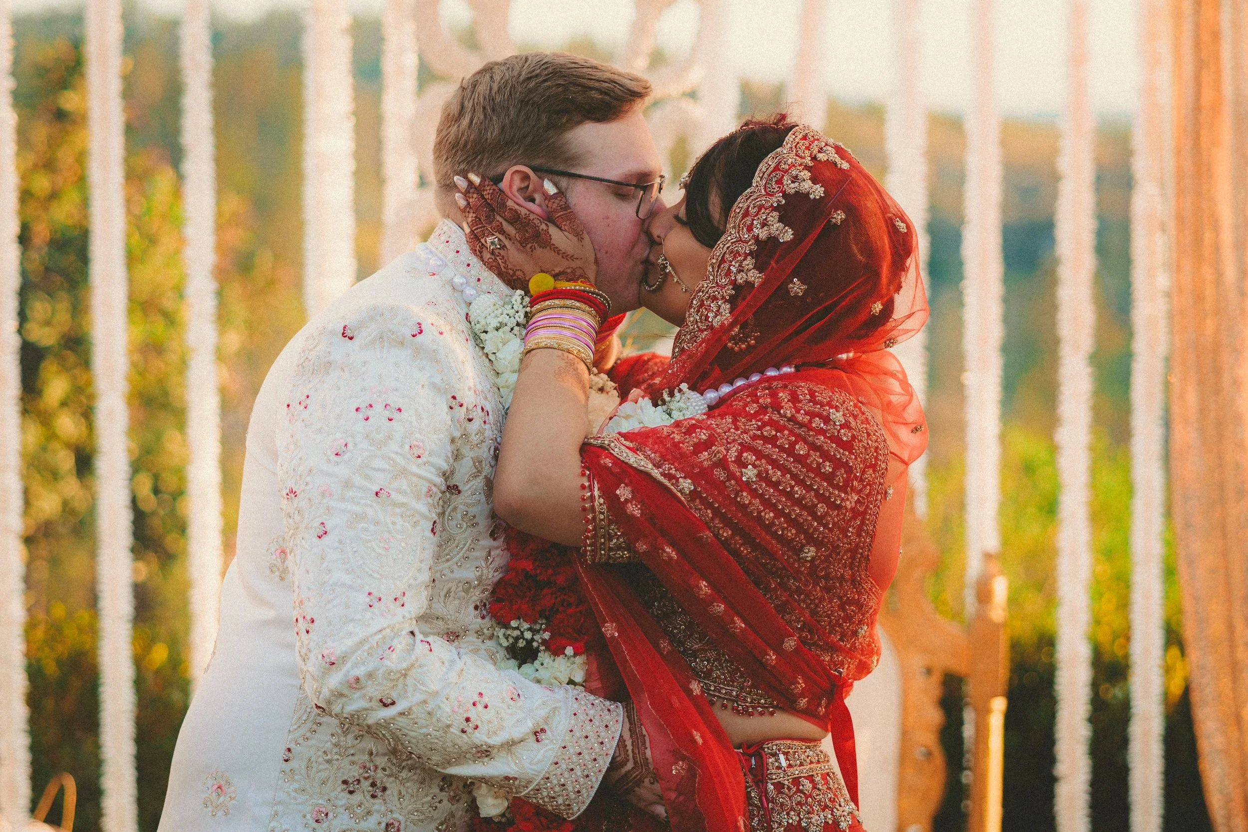 A couple dressed in traditional Indian wedding attire sharing a kiss, with trees and a wooden fence in the background.