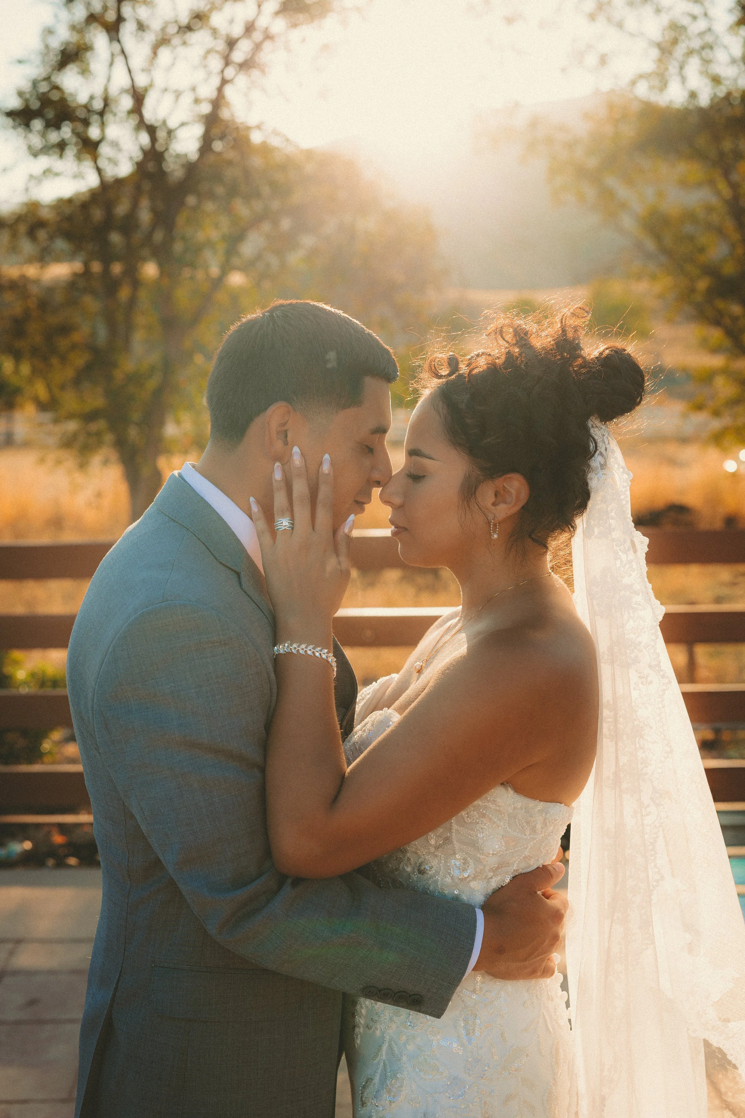 A bride and groom embrace outdoors during sunset, with trees and a wooden fence in the background.