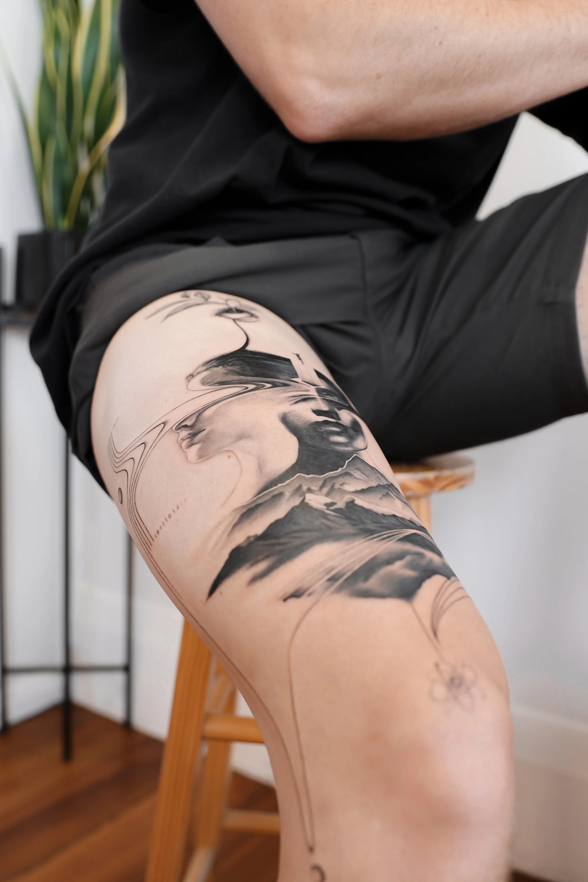 A person's thigh with a large black and gray tattoo featuring a woman's face, mountains, and abstract lines.
