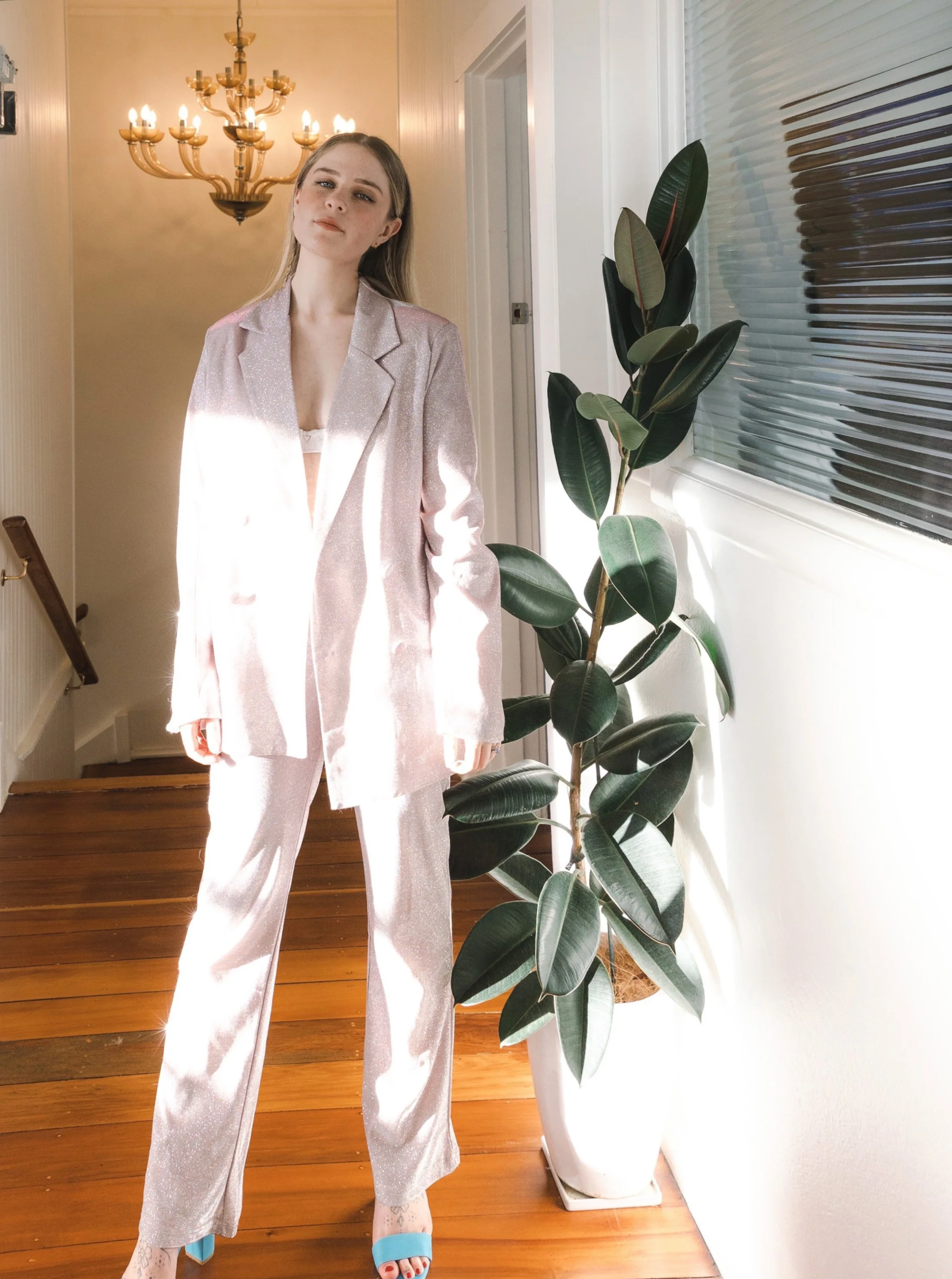 A woman in a shiny, light pink pantsuit standing indoors near a large potted plant with green leaves, illuminated by natural sunlight and a chandelier in the background.