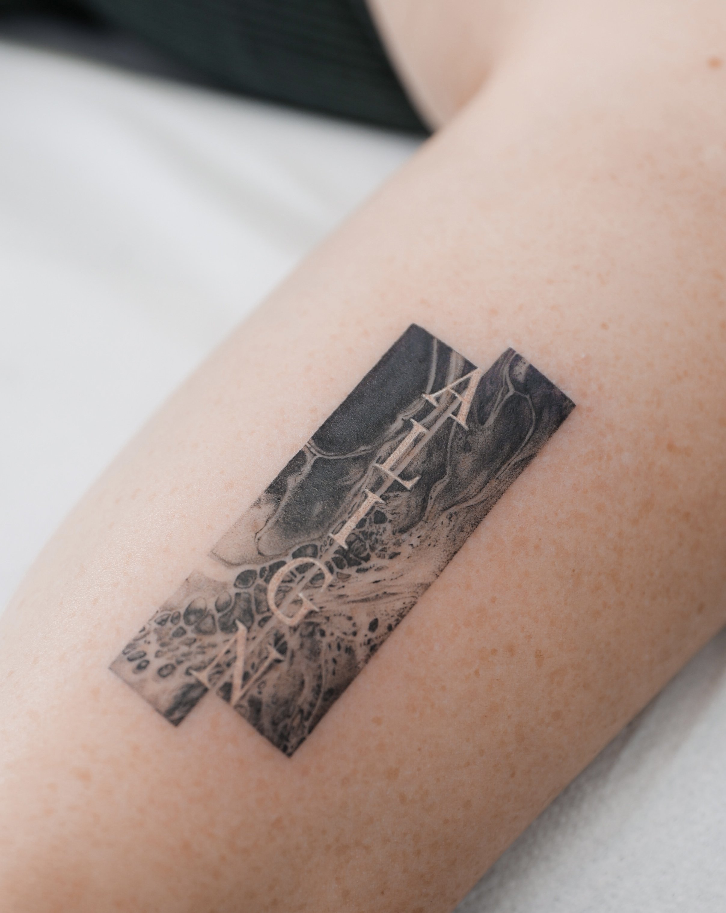 Close-up of a black and grey tattoo on a person's upper arm, featuring a landscape scene and abstract designs.