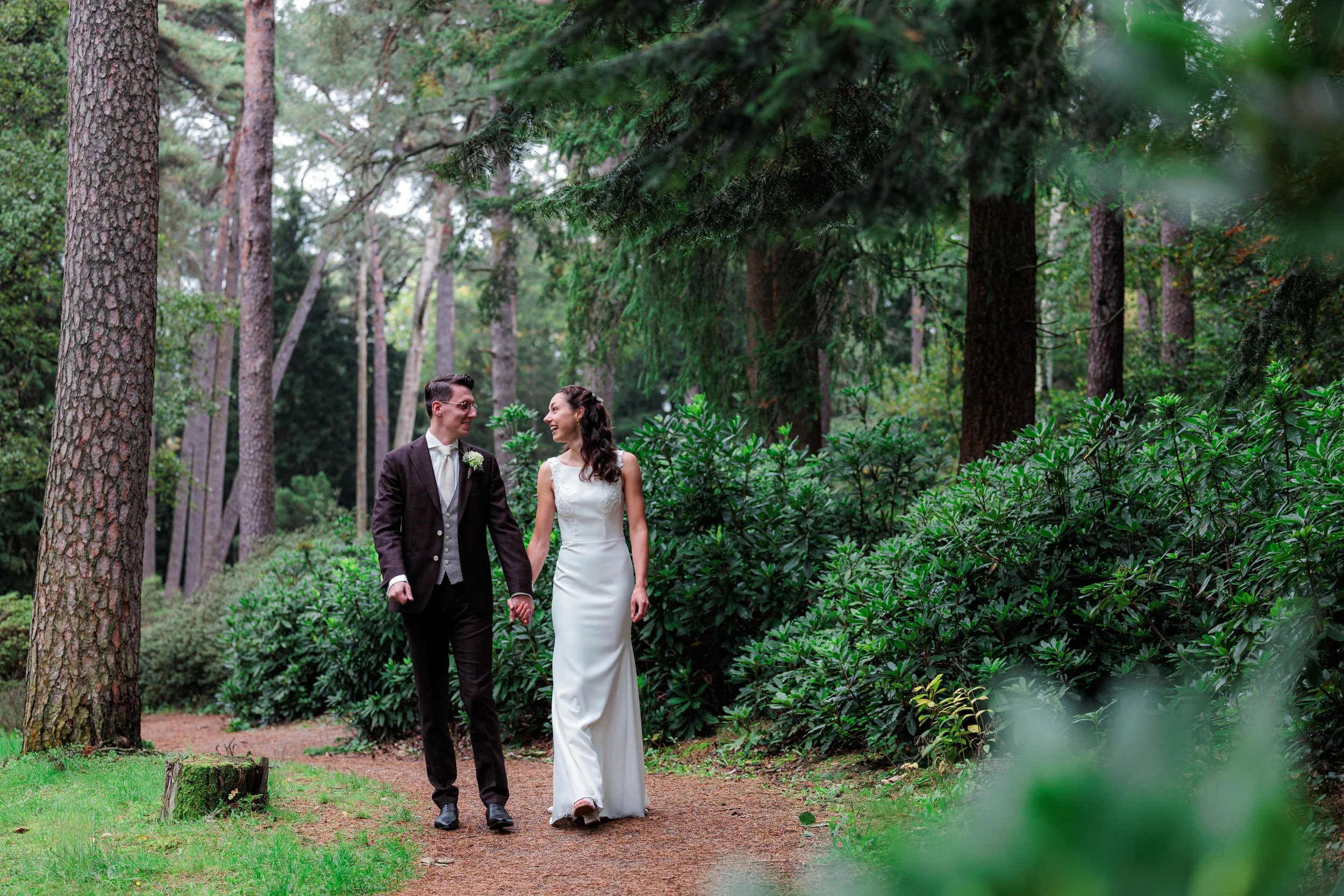 Een pas getrouwde paar wandelt hand in hand door een bos, zij lachend en kussend, omgeven door groene bomen en struiken.