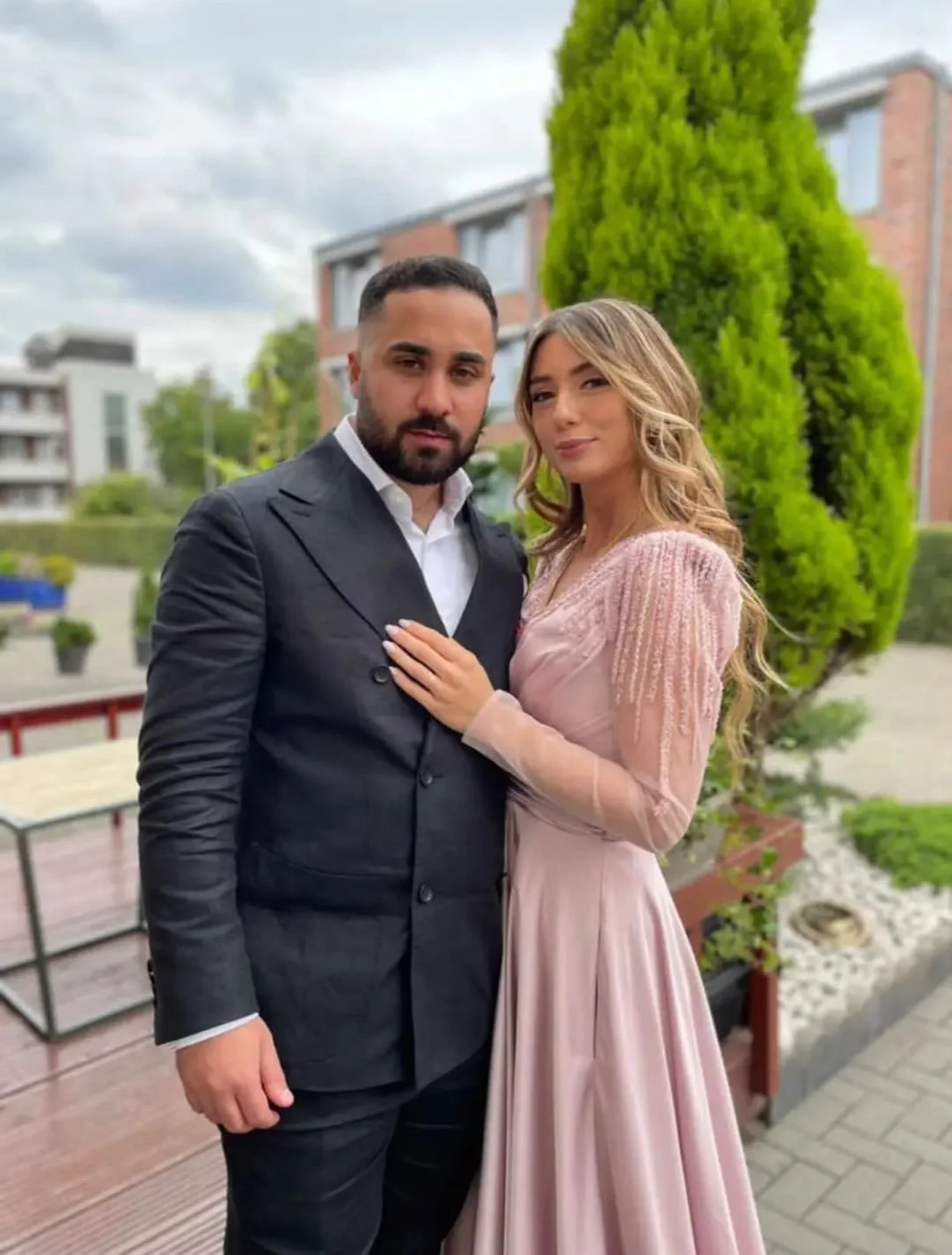 Ein Mann und eine Frau stehen nah beieinander vor einem Garten mit großen, grünen Bäumen, im freiem. Der Mann trägt einen schwarzen Anzug, die Frau ein langes, pinkfarbenes Kleid.