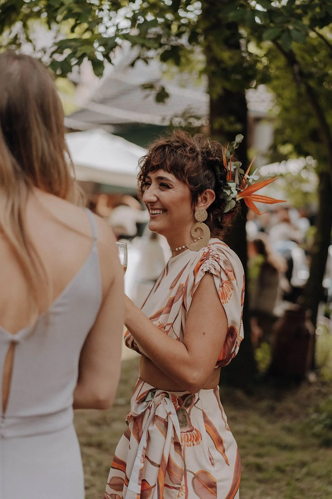 Eine lächelnde Frau mit kurzen, lockigen Haaren, großen Ohrringen und einem Blumen-Accessoire im Haar, trägt ein sommerliches Kleid mit Blattmuster bei einem Outdoor-Event.