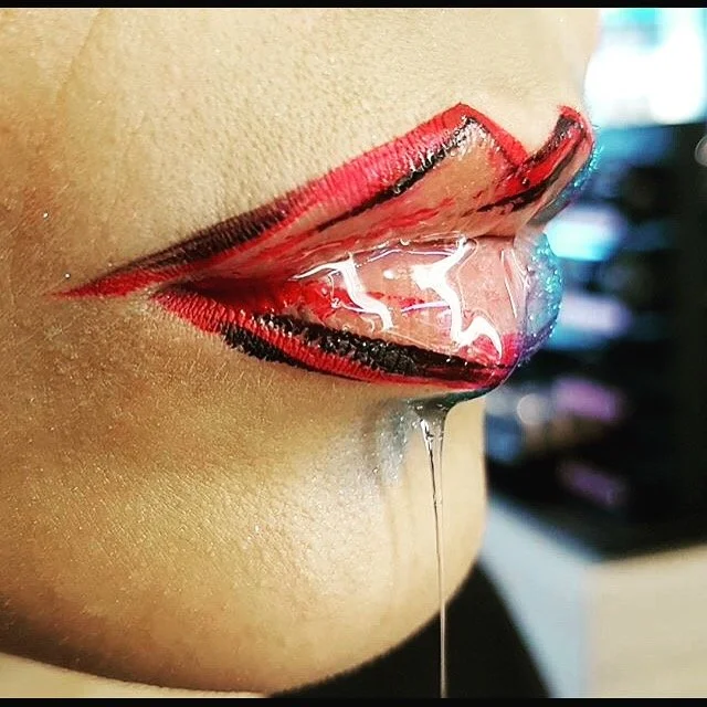 Ein künstlich gestalteter Mund mit roten und schwarzen Lippenstift und einer Zunge, die aus dem Mund herausragt, mit einer Flüssigkeit, die vom Mund herunterläuft.