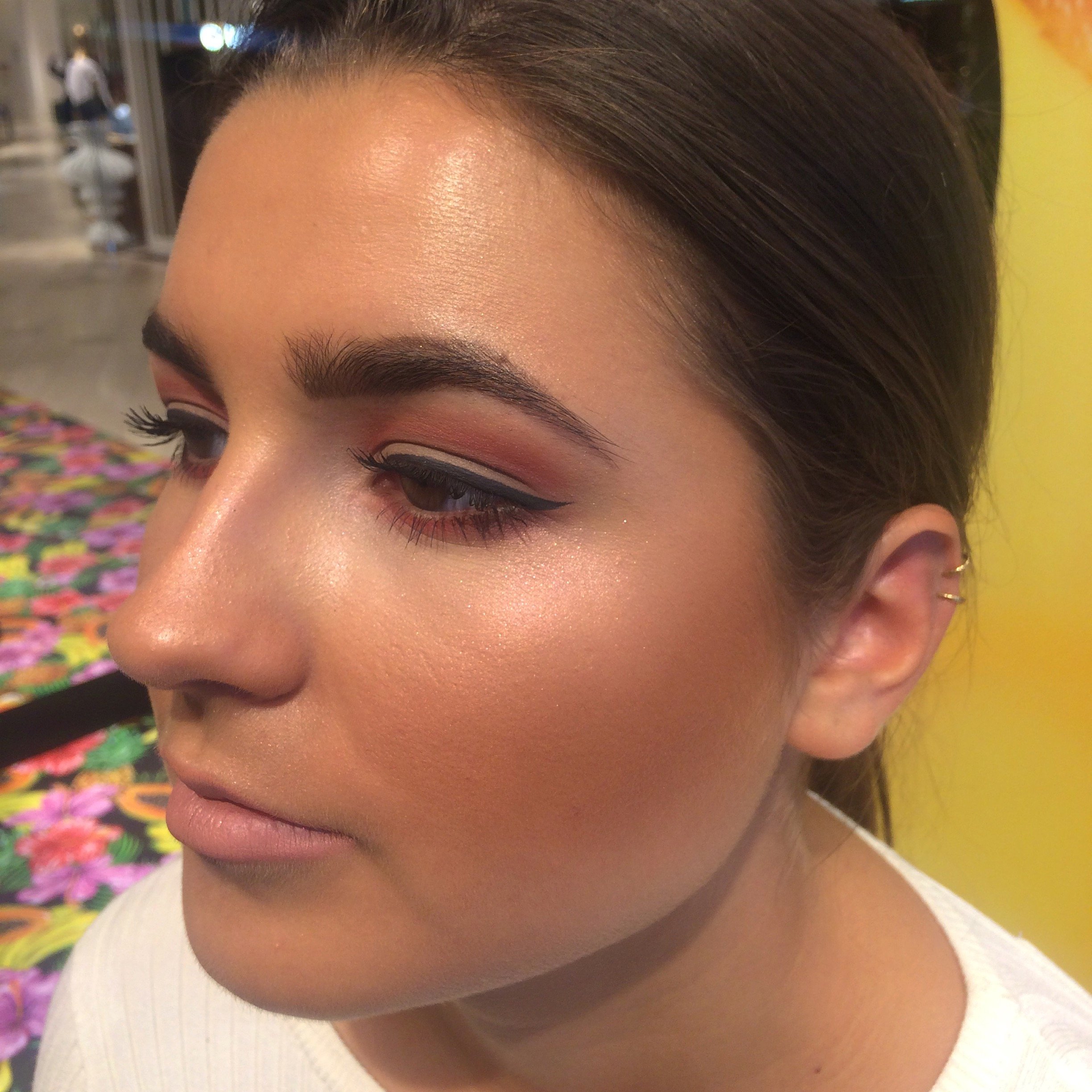 Nahaufnahme eines Gesichts mit glamourösem Make-up, dunklen Augenbrauen, auffälligem Eyeliner und glowy Haut, vor einem bunten Hintergrund.