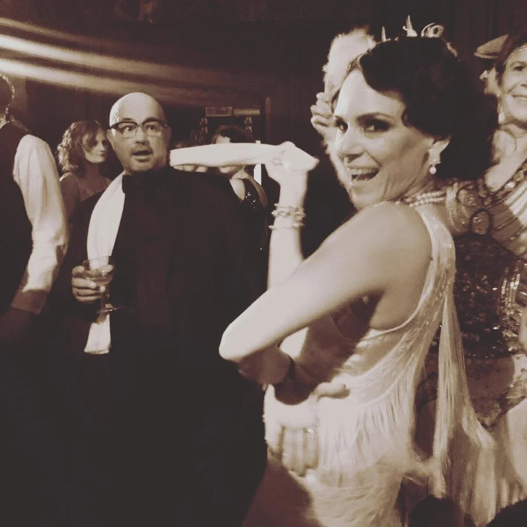 Menschen beim satten Fest in den 1920er Jahren, ein Mann hält ein Getränk in der Hand, eine Frau trägt ein elegantes Kleid, die Szene wirkt wie eine Feier aus dem frühen 20. Jahrhundert.