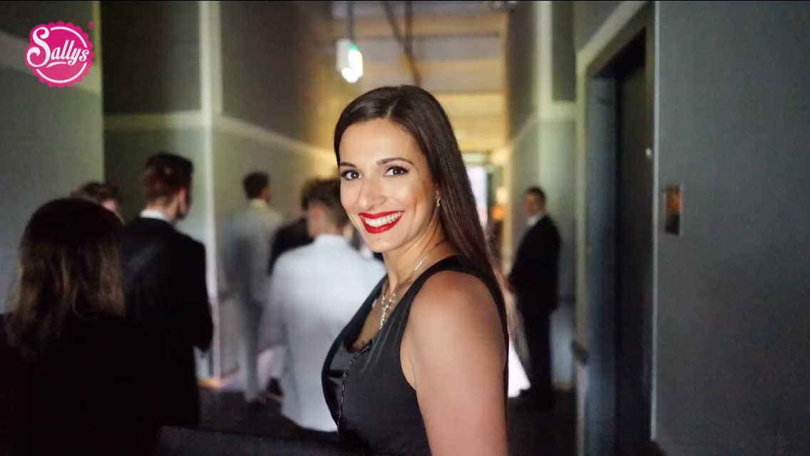 Eine lächelnde Frau mit dunklen Haaren und rotem Lippenstift, die in einem Hotel oder Veranstaltungsraum steht, im Hintergrund mehrere Personen in formeller Kleidung.