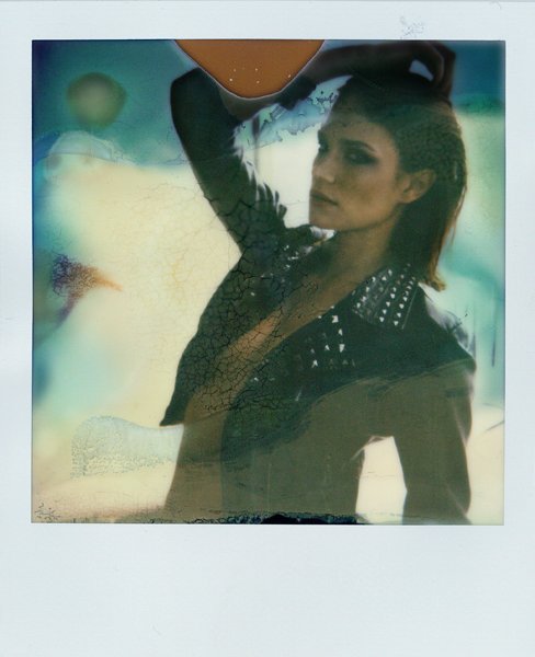 Eine Frau mit dunklem Make-up und einem schwarzen Oberteil mit Nieten, die in einem Polaroid-Foto posiert, während sie einen Bikini über einem Schulter trägt.