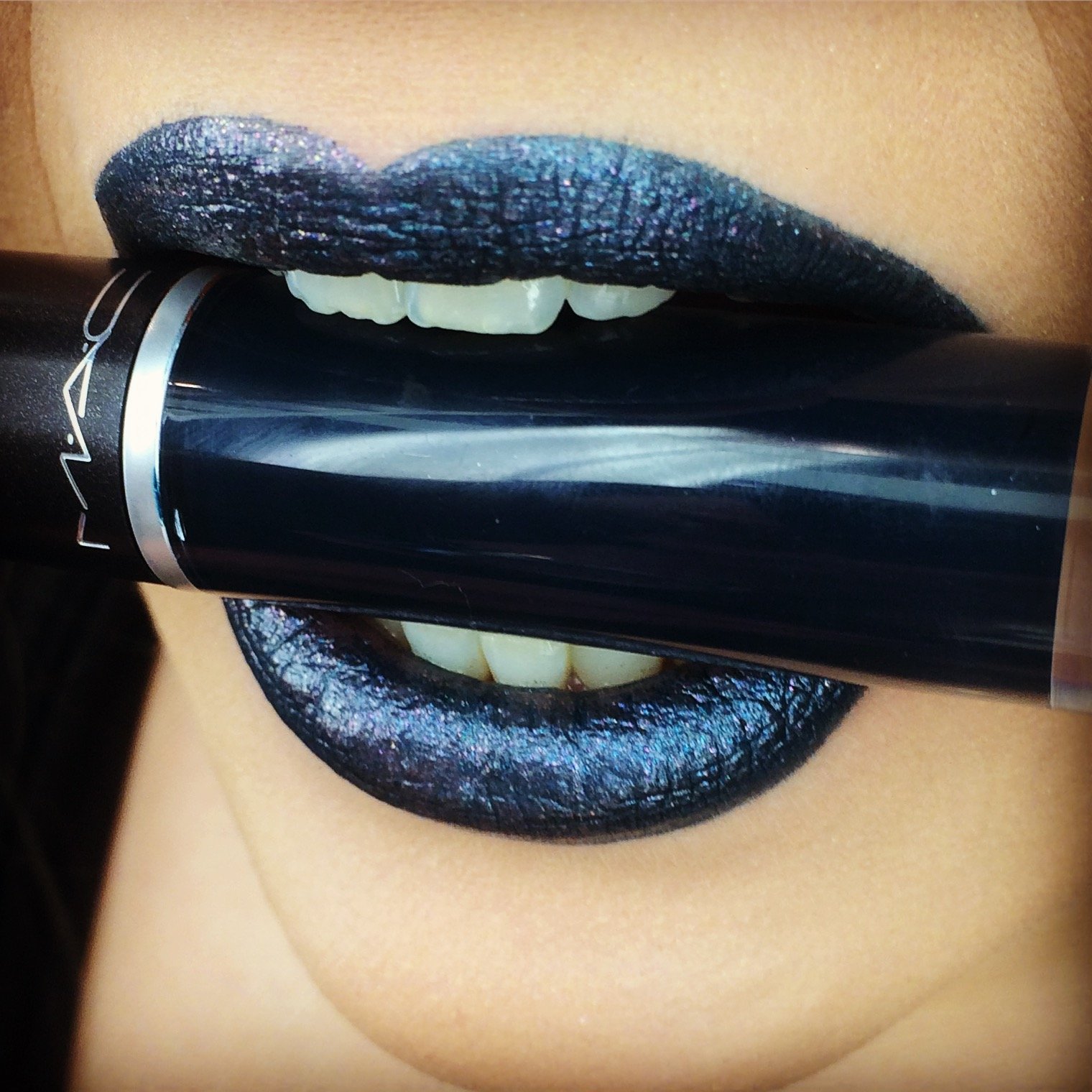 Nahaufnahme eines Mundes mit schwarzen, schimmernden Lippenstift, der eine Lippenstiftverpackung zwischen den Zähnen hält.