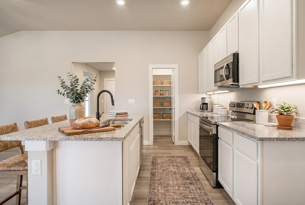 Modern kitchen with white cabinets, granite countertops, stainless steel oven and microwave, coffee maker, potted plant, bread on a cutting board, and a hallway with pantry shelves.