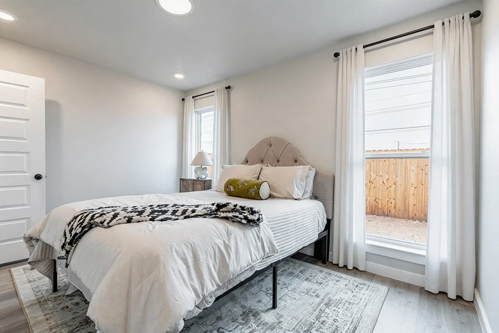 A bedroom with white walls, a bed with a beige upholstered headboard, white bedding, and decorative pillows, including a green pillow, a nightstand with a lamp, and white curtains on two windows with a view of a wooden fence outside.