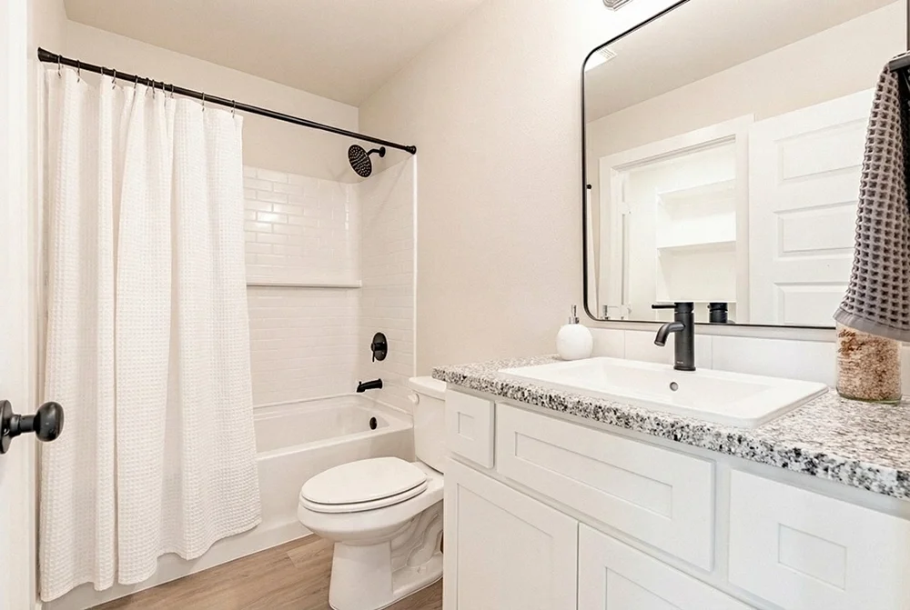 Bathroom with a bathtub and shower, white tile surround, white shower curtain, toilet, granite countertop with a sink, large mirror, and various bathroom accessories.