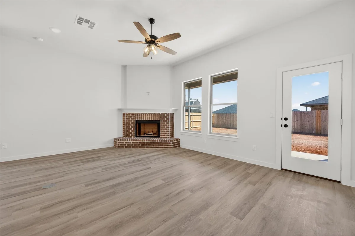 Empty living room with white walls, wood floor, brick fireplace, ceiling fan, two windows, and a door leading outside.
