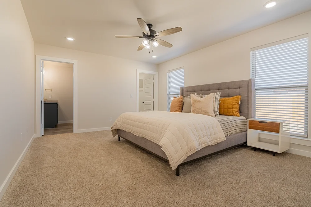 Spacious bedroom with beige carpet, large bed with gray upholstered headboard, multiple pillows, two windows with blinds, ceiling fan, and an open door to a small bathroom.