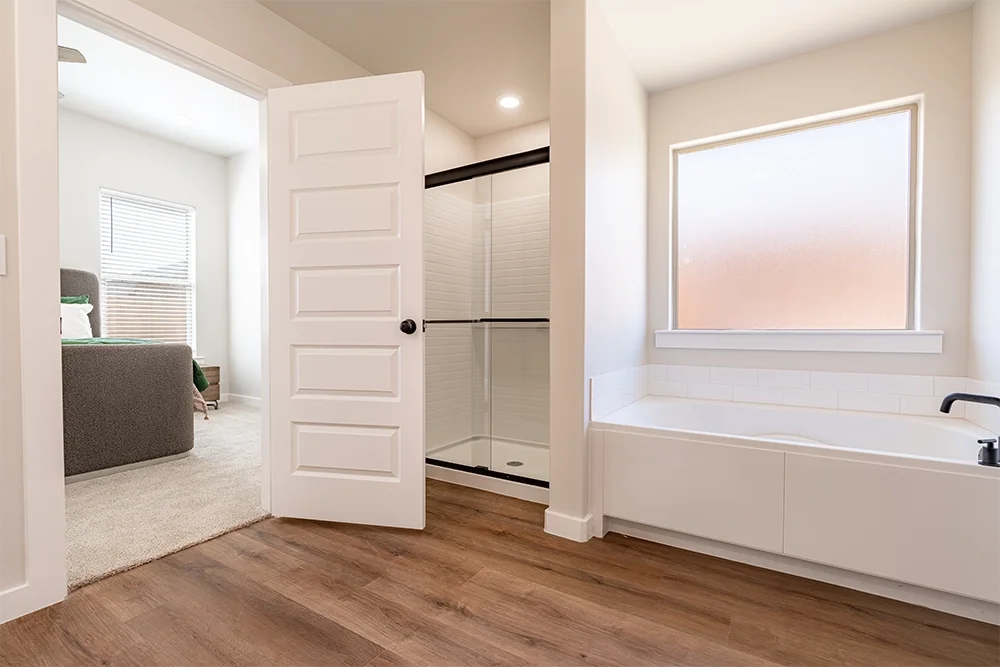 Bathroom with a bathtub under a frosted window, a walk-in shower with glass doors, and a sliding door to a bedroom with a bed and window.