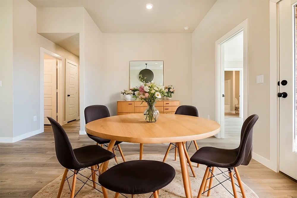 A dining room with a round wooden table, four black cushioned chairs, a glass vase with flowers, a wooden sideboard with a mirror above it, and a doorway leading to another room. Light-colored walls and wood flooring are visible.