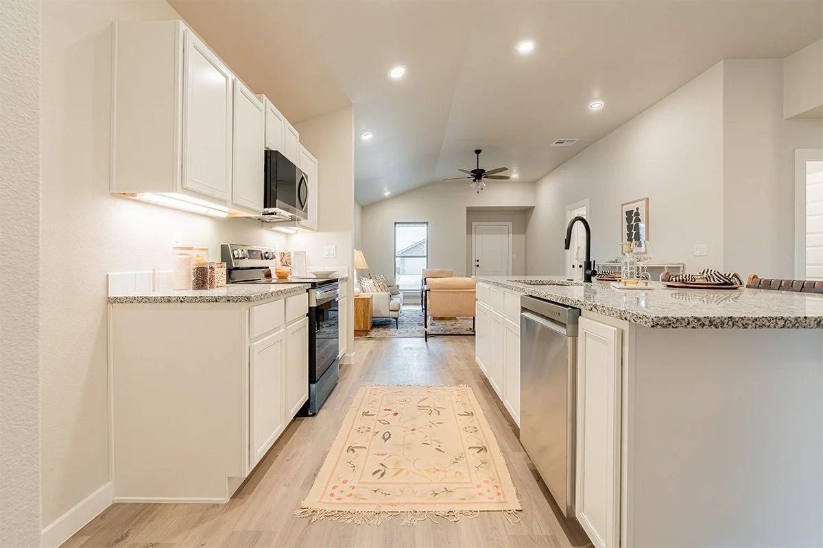 Modern kitchen with white cabinets, granite countertops, stainless steel oven and microwave, and a bar seating area. The kitchen opens to a living room with beige sofas and a small rug, with natural light coming from a window and door in the backgrou