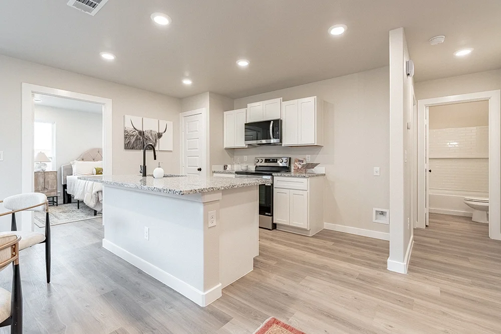 Open-concept kitchen with white cabinets, granite countertops, stainless steel appliances, and a breakfast bar. Adjacent to a bedroom with a bed and side tables, and a bathroom with a toilet and white tiles.