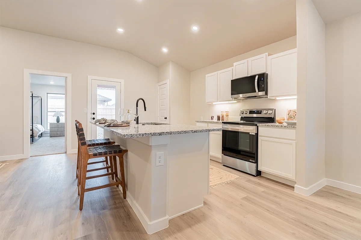 Modern kitchen with white cabinets, stainless steel appliances, granite countertops, and a breakfast bar with wooden chairs, overlooking a dining area with a bedroom visible through an open door.