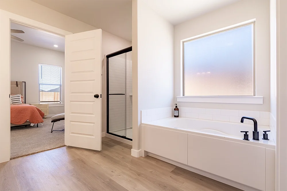 A modern bathroom with a large frosted window, white bathtub with black faucet, and a bottle of soap. Adjacent bedroom with a bed, window, and beige carpet.