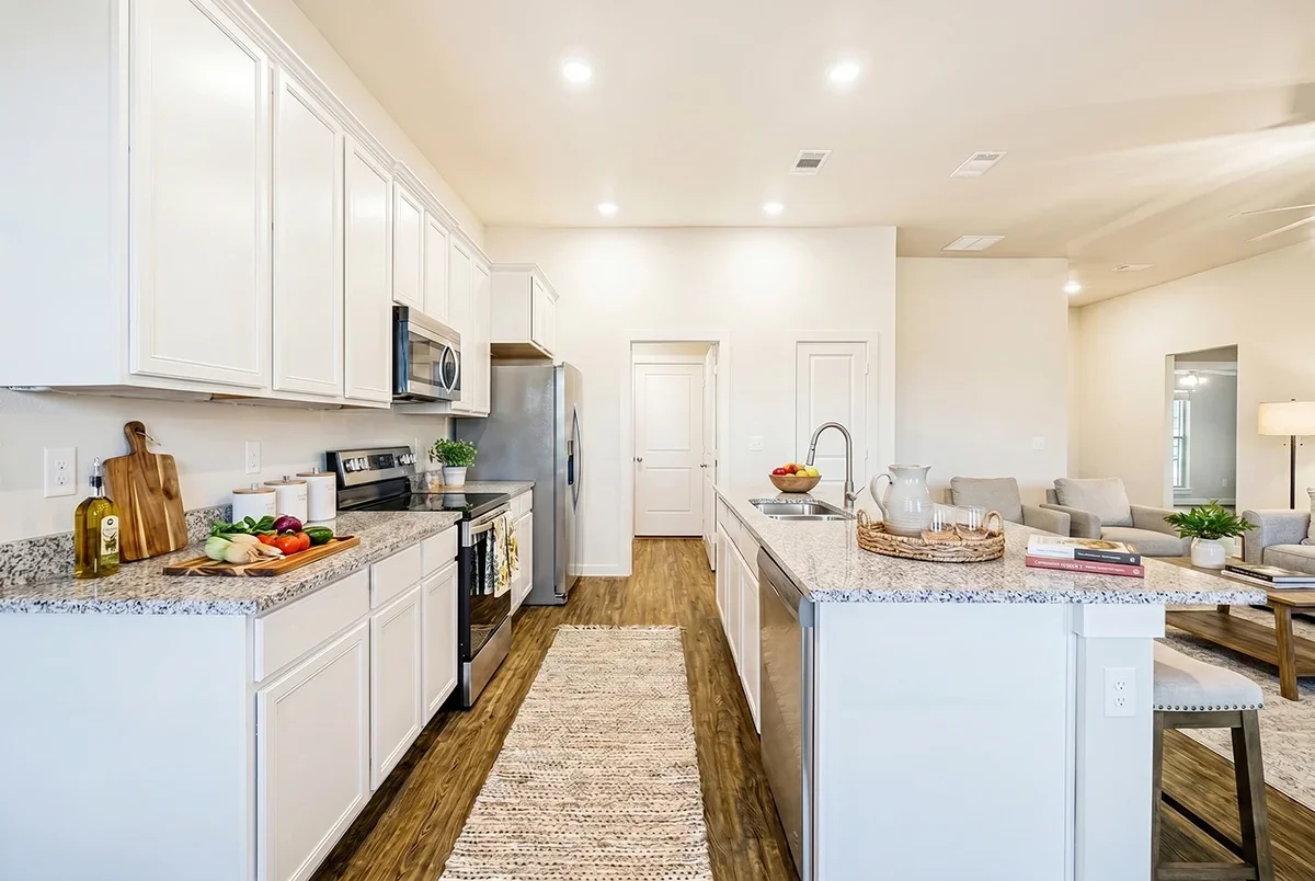Bright modern kitchen with white cabinets, granite countertops, stainless steel appliances, and hardwood floors, adjacent to a living area with beige seating.