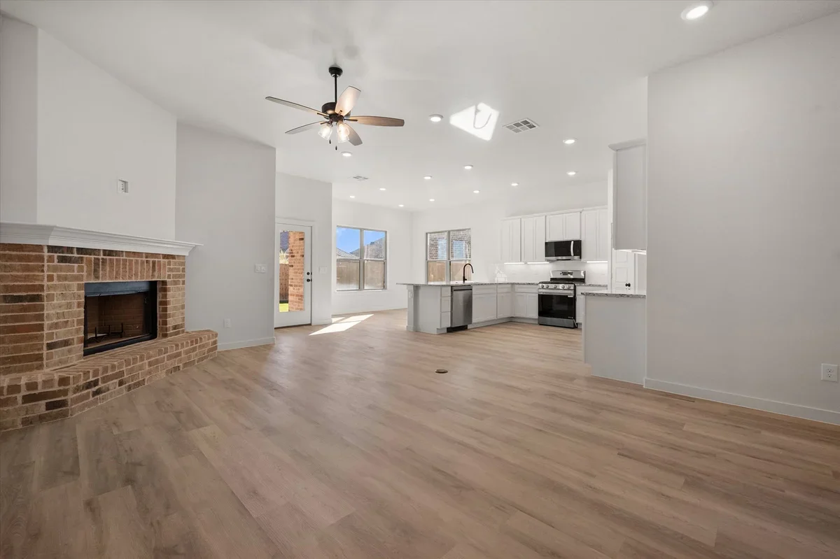 Empty living room with brick fireplace, ceiling fan, and open kitchen with white cabinets, black appliances, and wooden floors.