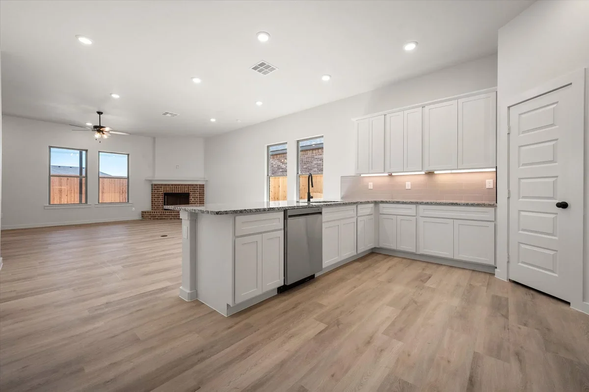 Open-concept living room and kitchen with white cabinets, granite countertops, hardwood floors, and a brick fireplace.