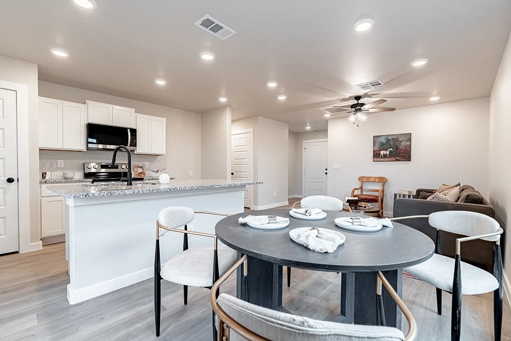 Open-concept kitchen and dining area with white cabinets, granite countertops, stainless steel appliances, a black dining table set with white placemats and napkins, and a seating area with a sofa, armchair, and wall art.