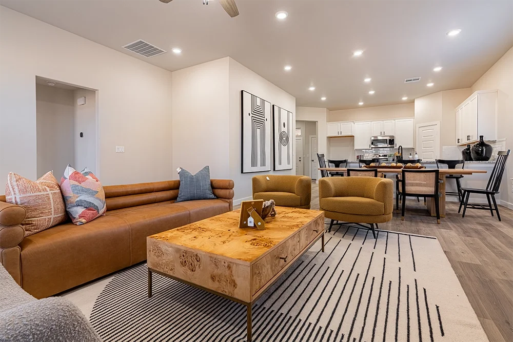Open-concept living and kitchen area with a brown leather sofa, yellow armchairs, a wooden coffee table, abstract art on the wall, and white cabinetry in the kitchen.