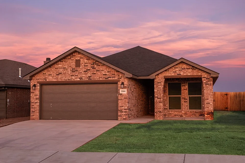 A brick house with a garage and front lawn during sunset, with a pink and purple sky.