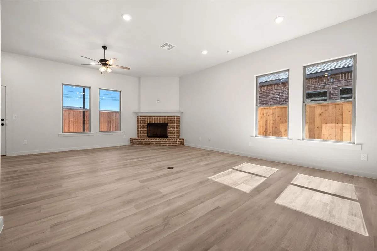 Empty living room with light-colored wooden floors, white walls, a brick fireplace, four large windows, a ceiling fan, and recessed lighting.