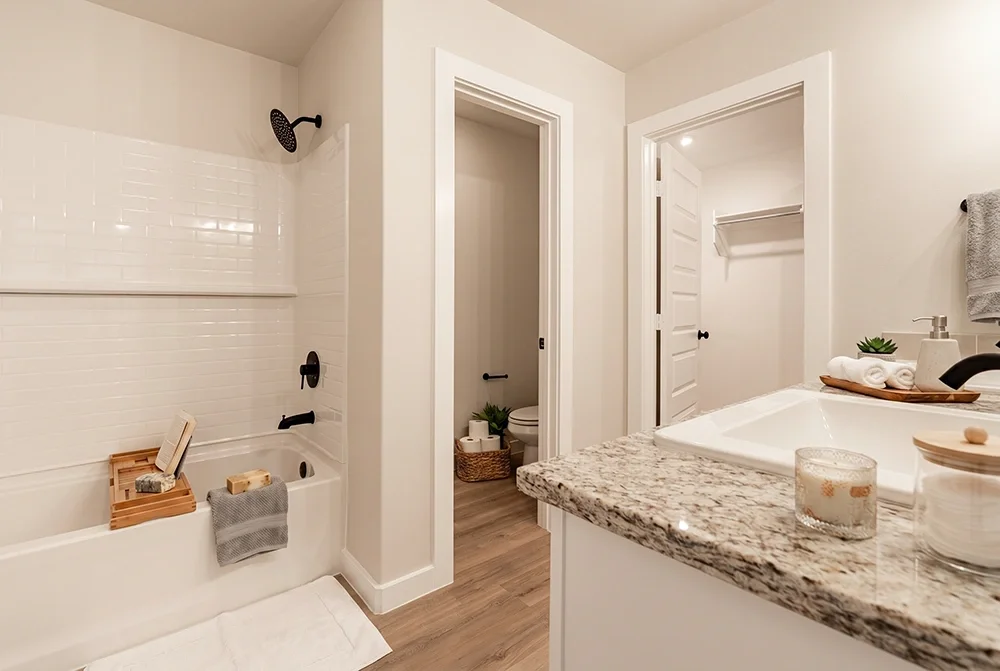 Modern bathroom with white tiled shower, granite countertop sink, and a separate toilet area. Decor includes towels, a soap dispenser, and small plants.